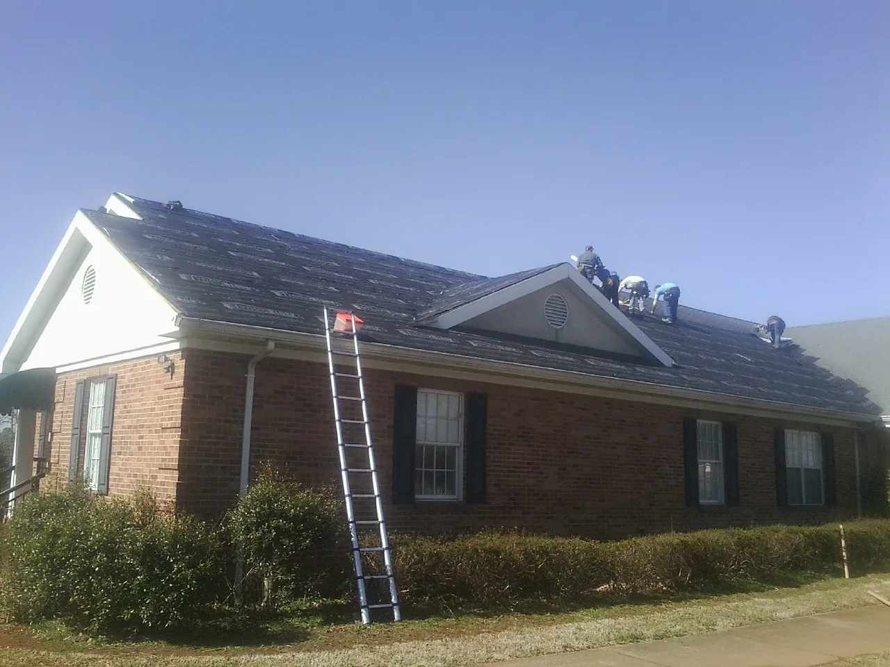 Crew tearing shingles off a brick ranch roof with a ladder set — Metal Roof Repair work by our crew in Ackworth
