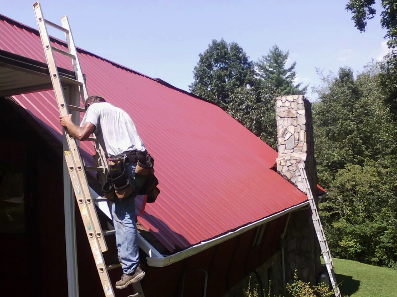 Roofer on a ladder beside a red metal roof and stone chimney — Soffit & Fascia Repair by Roofing Brothers in Ackworth